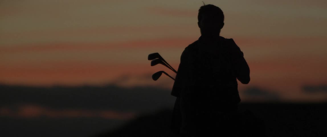 Man carrying golf clubs is standing outside after sunset