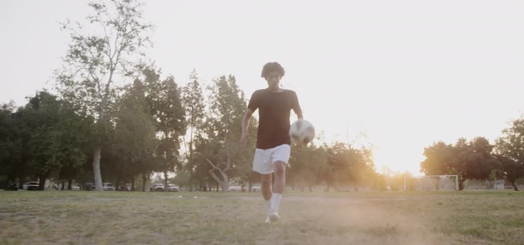Boy is kicking a soccer ball in an open soccer field as sun sets
