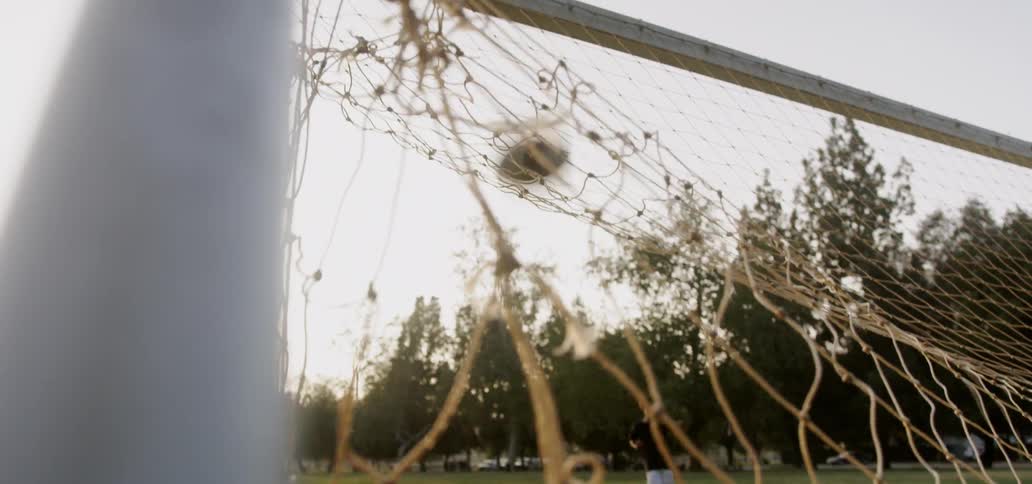 Boy kicks soccer ball into goal post as sun sets