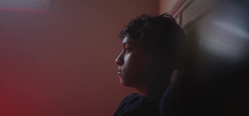 Teenage hispanic person leaning against a headboard and shaking their head slowly back and forth