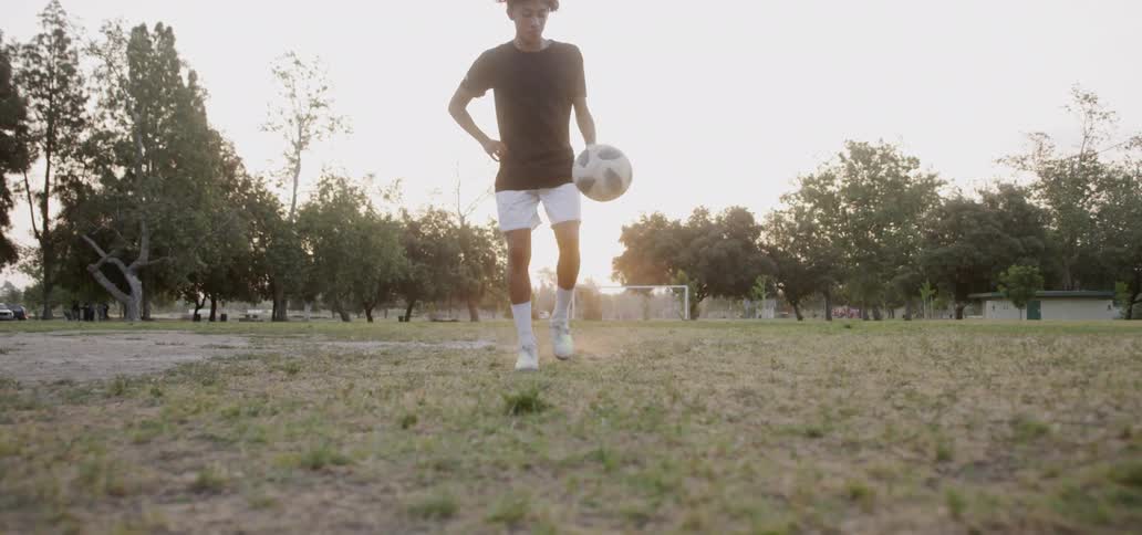 Boy is kicking soccer ball into the air on field during sunset