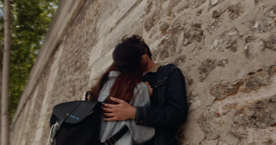 A woman kisses a man leaning against a wall on the sidewalk