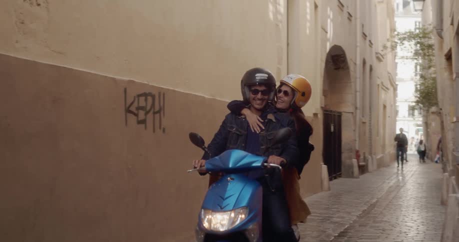 A young couple on vacation ride on a scooter through cobblestone alley in Europe