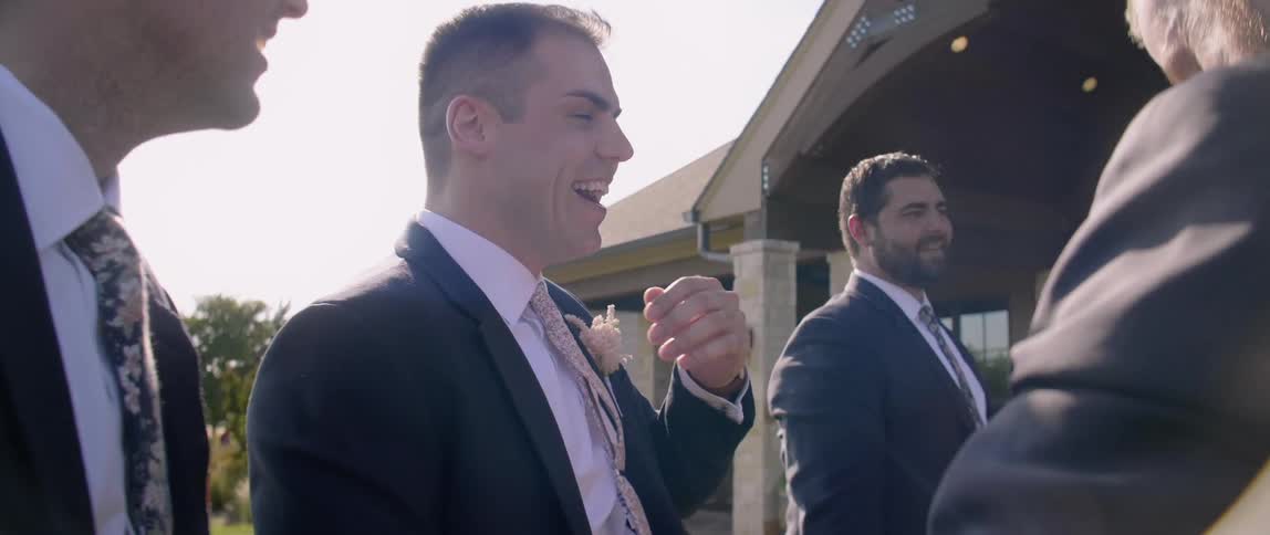A groom and groomsmen laugh outside on a sunny day