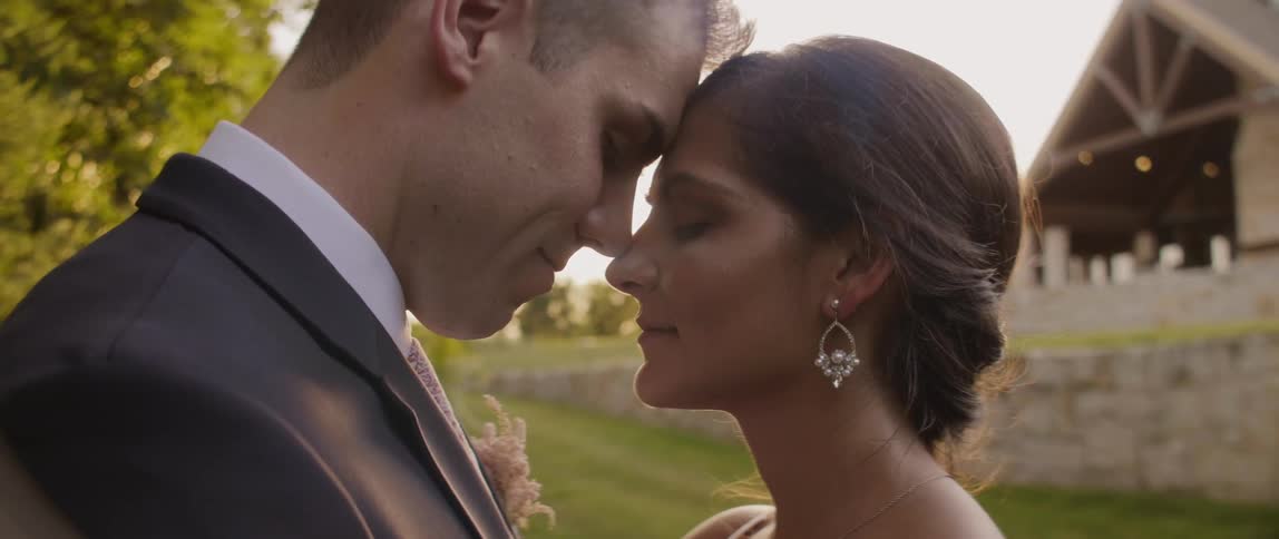 A bride and groom are standing nose to nose outside in daylight