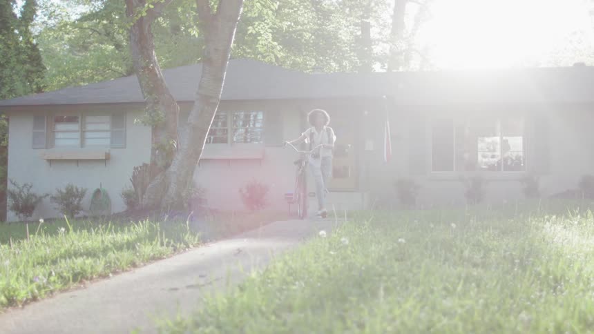 Woman walks her bike down a sidewalk outside of a house on a sunny day