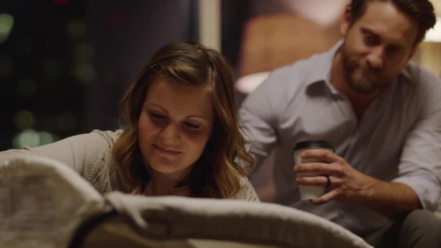 A man is patting his partner's back as she tends their baby inside at night