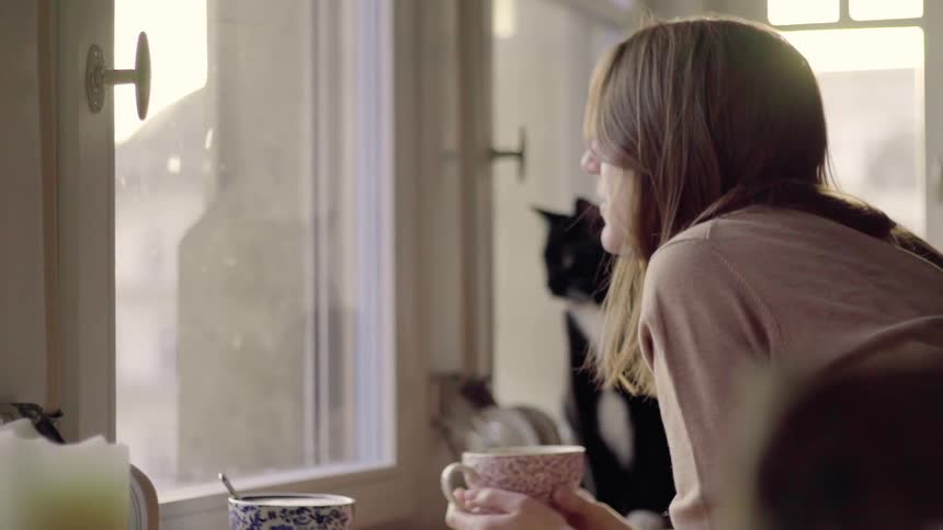 Woman holds a mug and brushes her hair back while a cat sits on a table