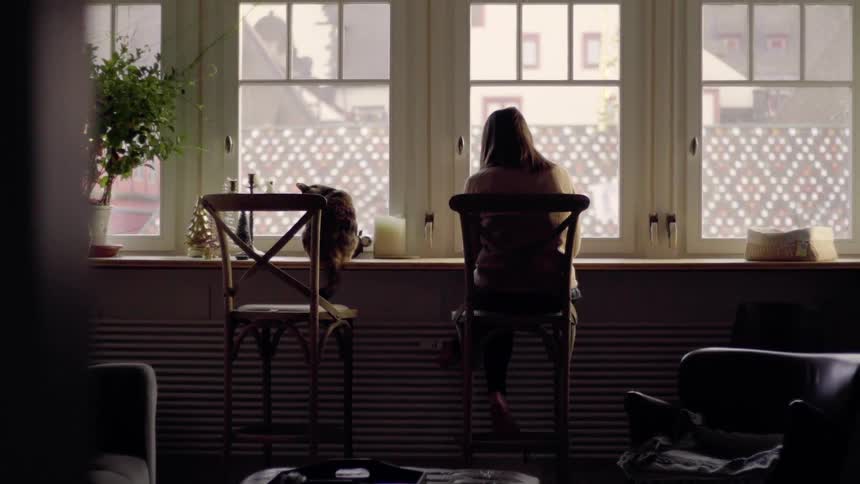 A woman sits in a chair by the windows with a cat