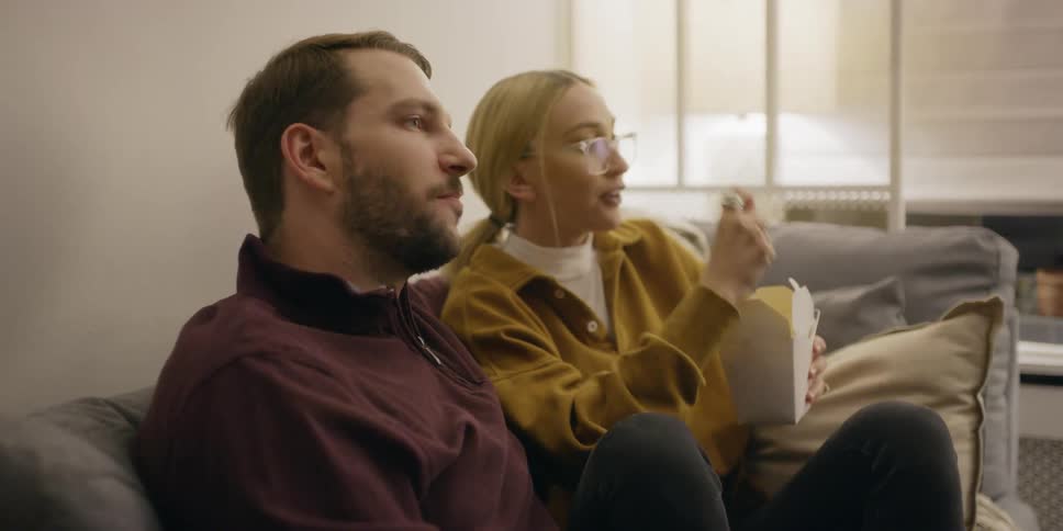 A man and woman eat together on the couch