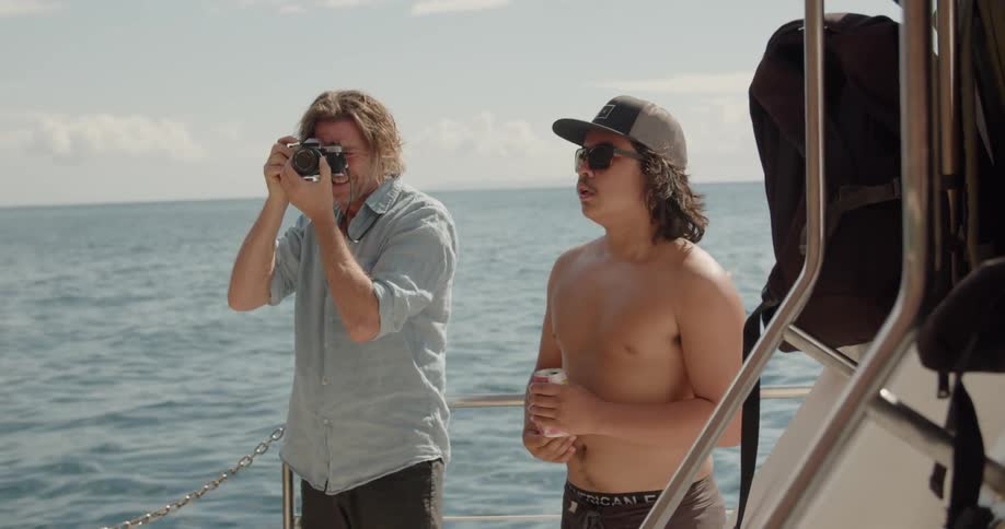 Two men talk on the back of a boat as one takes a photo