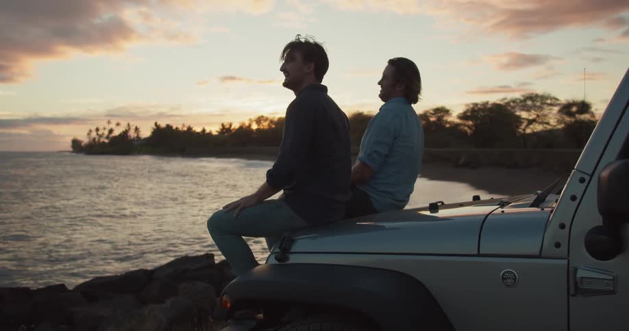 Two men sit on the hood of a vehicle near the lake