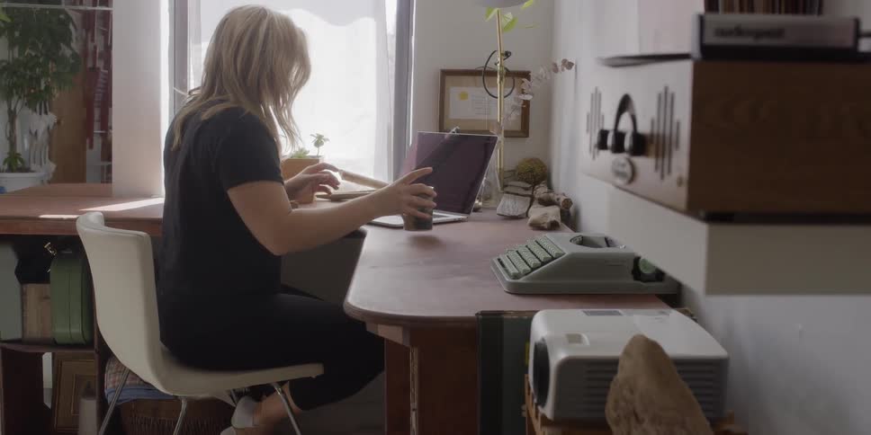 Woman sits down at a desk and takes a sip from a coffee mug