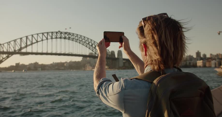 A man holds up a cellphone and films a bridge at sunset