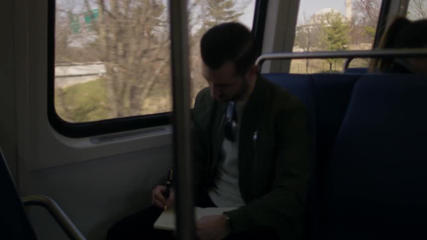 A man is sitting on a commuter train writing in a journal in daylight with monuments in background