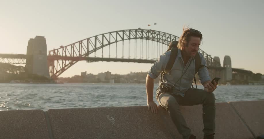 A man takes a photo sitting on a wall in front of a bridge and harbor at sunset