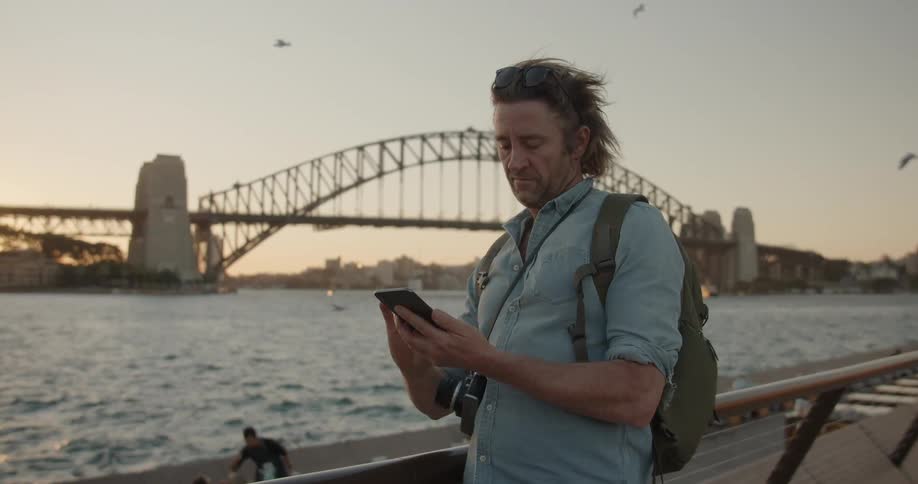 Man uses his phone next to a city river at sunset