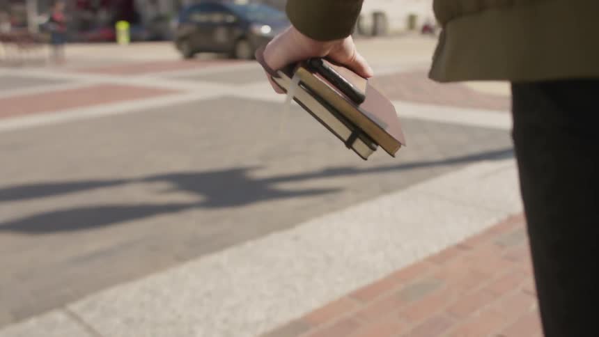 A pedestrian crosses a city street with books in hand