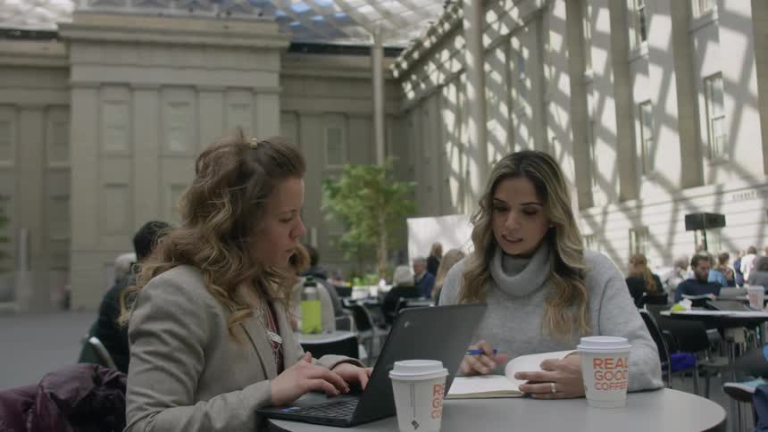 Two women sit at a table working on a laptop and looking at notes