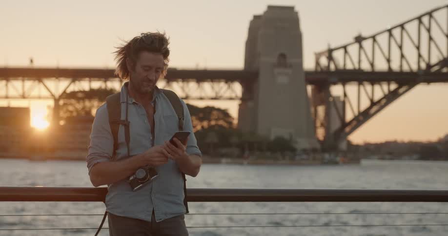 Man next to a river in a city typing on his phone at sunset with a bridge behind him