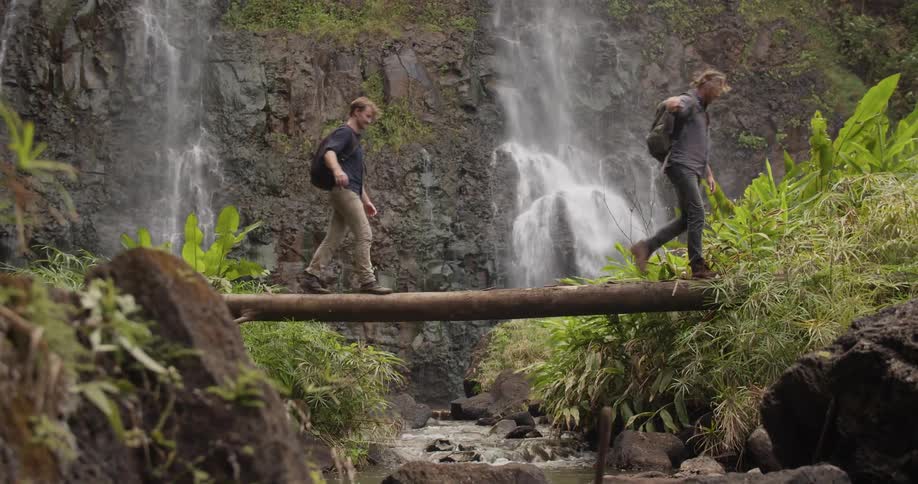 Two men walk across a log bridge near a waterfall