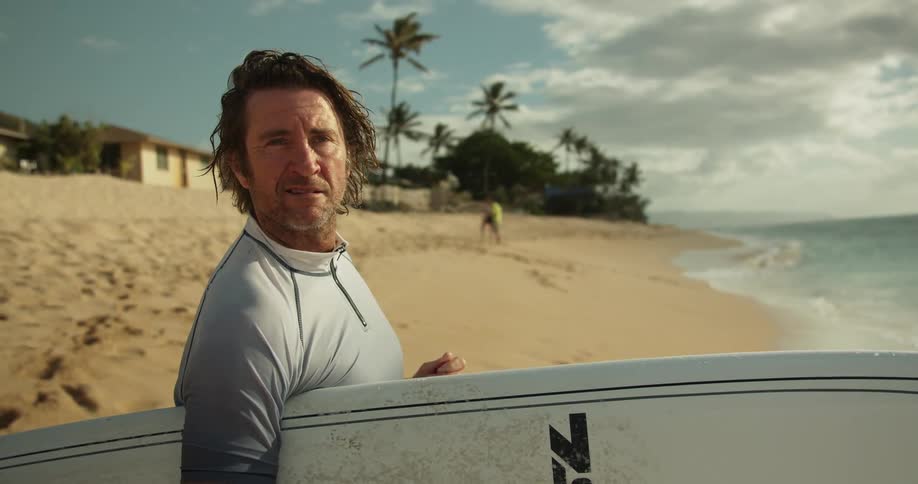 A man stands near the water with his surfboard