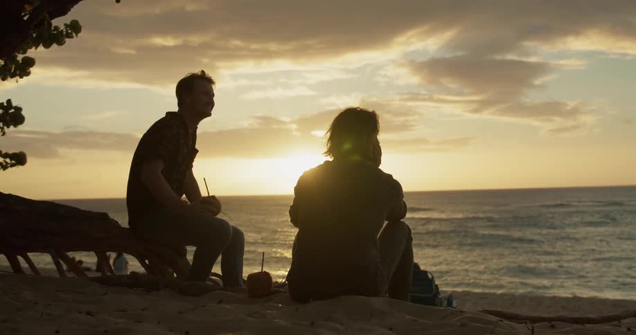 Two men are sitting on a beach at sunset having a drink and talking