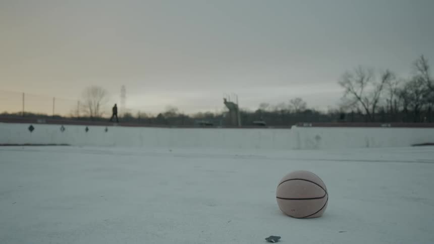 A basketball sits on the ground as a person walks past at sunset