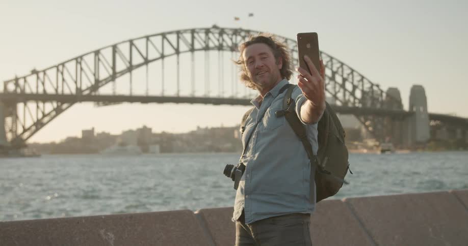 A man takes a selfie by the water and bridge