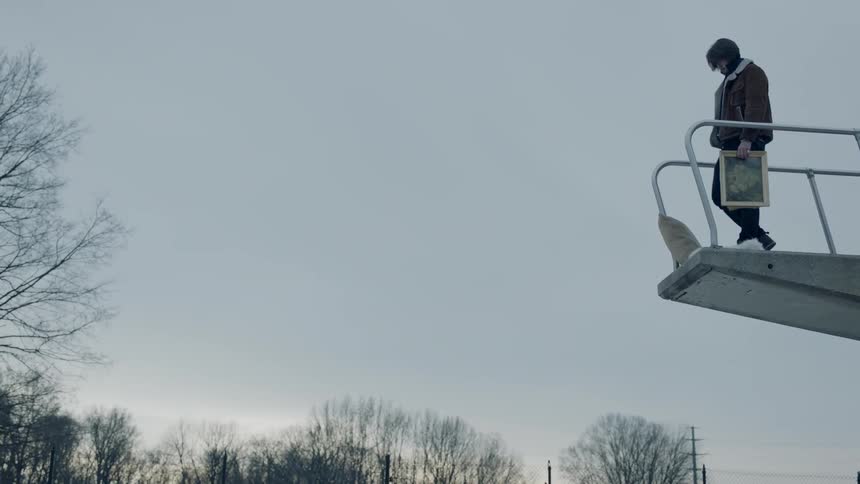 Man carrying a photo walks to the edge of a diving board and drops it