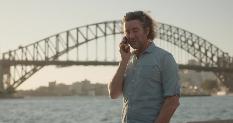 A man talks on the phone next to a city river at sunset