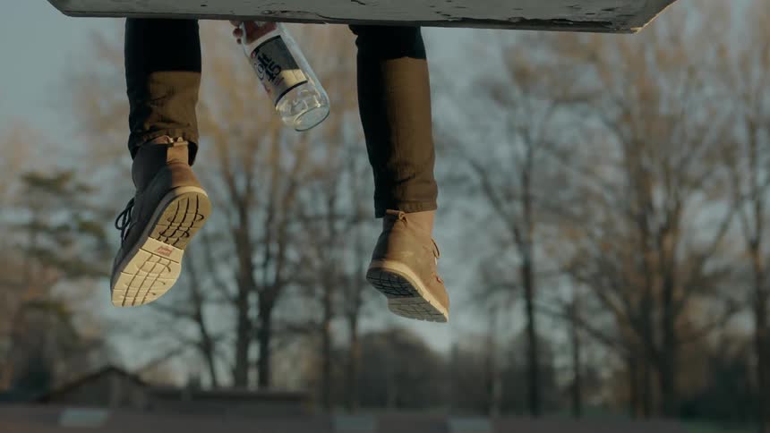 A person sits on a ledge outdoors swinging their legs and holding a bottle