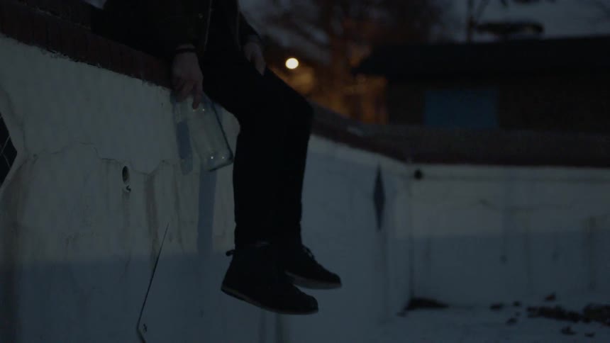 A person sitting on a wall breaks a glass bottle against it