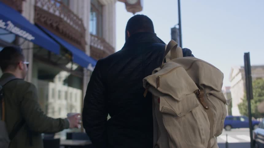 Two men are talking and walking on a city sidewalk in daylight with backpacks
