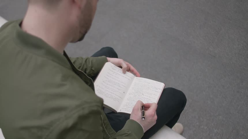 A man is sitting outside writing in a notebook in daylight