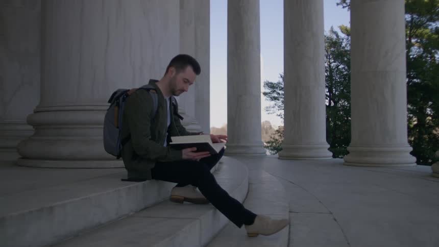 A man is reading on the steps of a monument on a sunny day