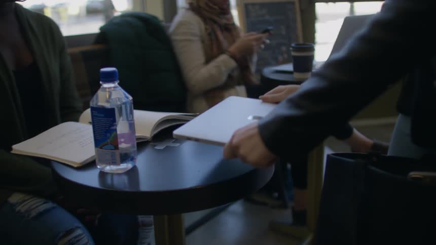 A woman pulls an apple laptop from a bag and puts it on a small table in a coffee shop