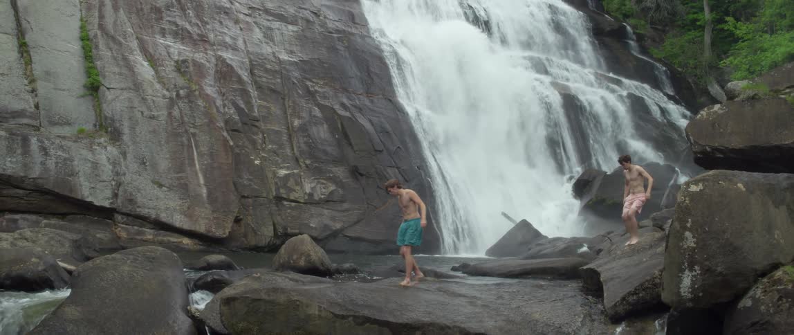 Two boys walk on rocks near a waterfall