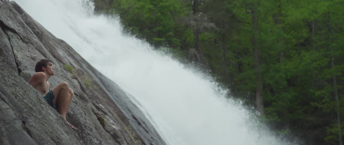 A mountain stream crashing down and over rocks while a man sits on a cliff side nearby