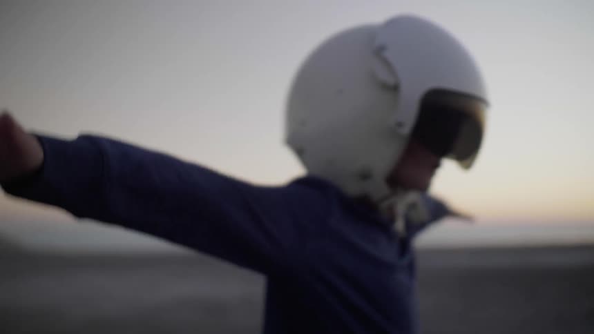 A young boy wearing an aviator helmet runs with his arms out as if he is flying