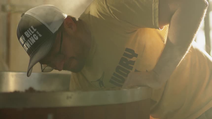 A man is scooping hops out of a brewing vat inside in daylight