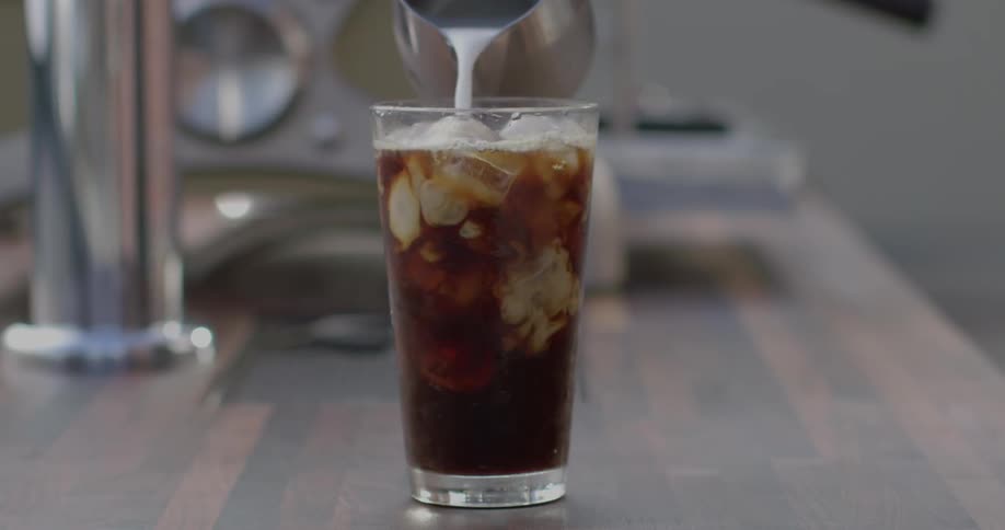 Cream pours from a pitcher into a glass of iced coffee