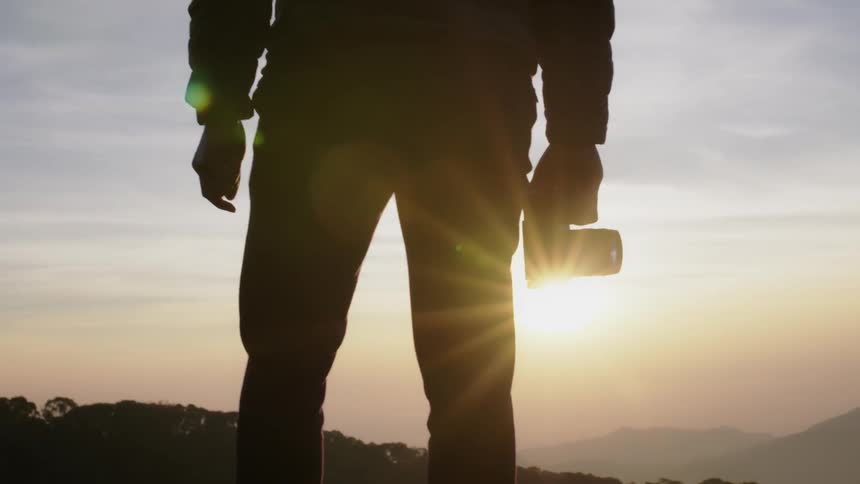 A man stands on a mountain holding a camera at dawn