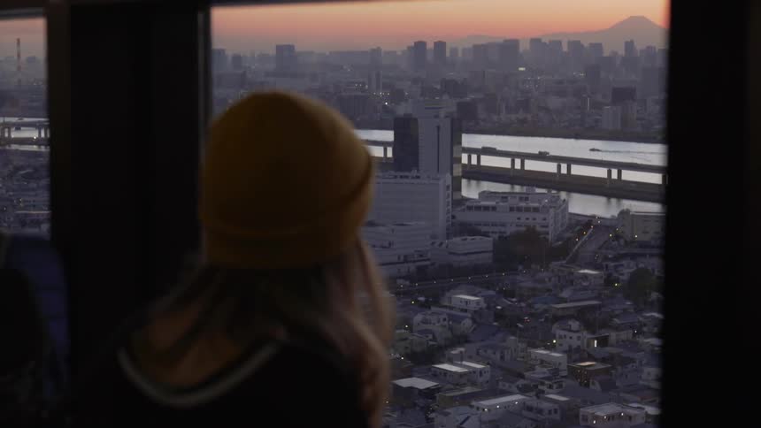 Young woman standing alone looks out from a high window toward downtown Tokyo