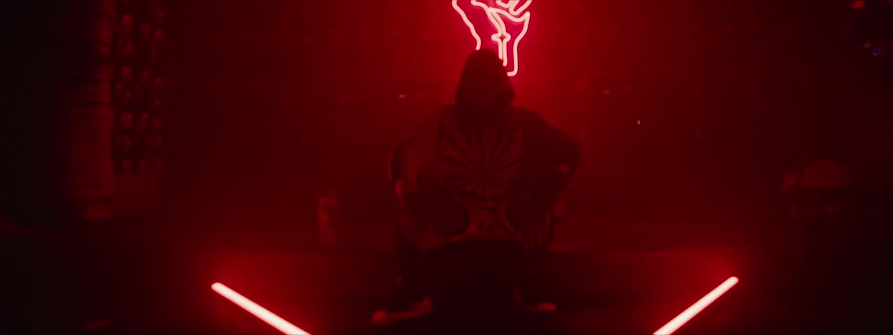 A man in a hood sits in a dark room with red flashing lights behind him