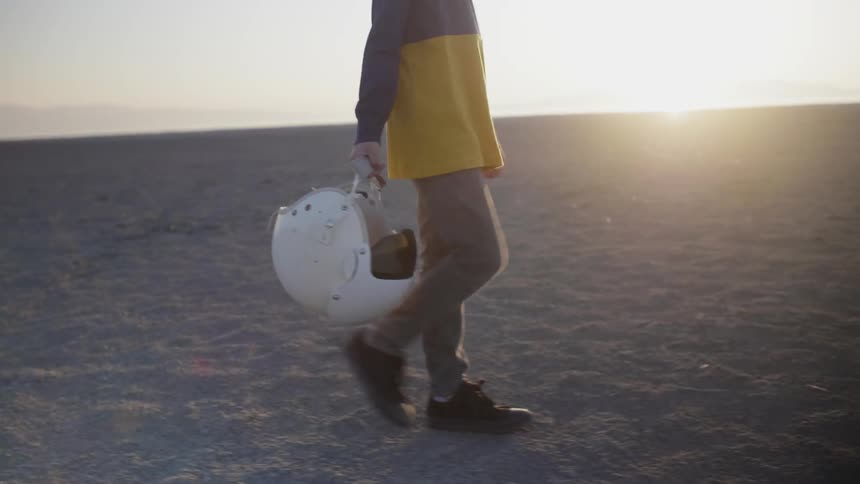 A boy holds a helmet while walking in a desert at sunset