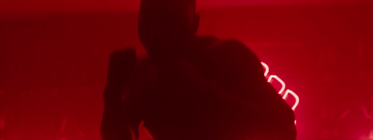 A man in a hoodie is shadow boxing in a red room with a red neon fist on the wall