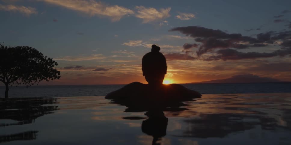 A woman standing in a pool located next to the ocean watching the sun as it sets