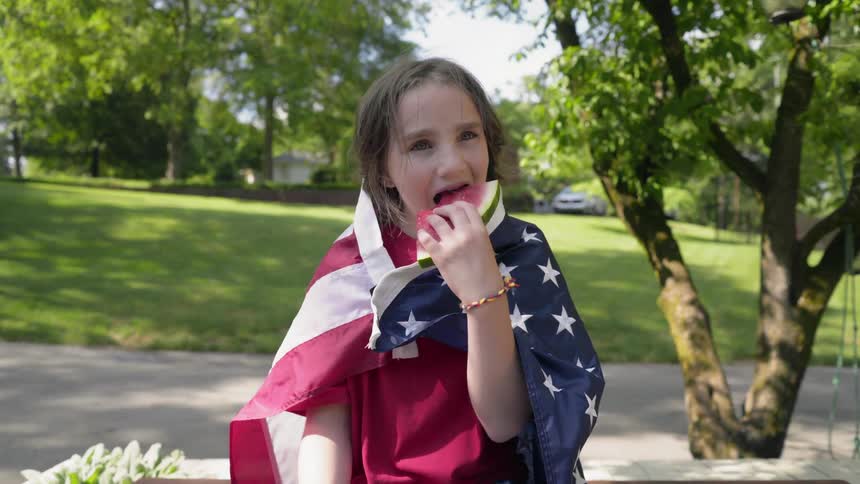 A young boy eats watermelon with an american flag draped around him