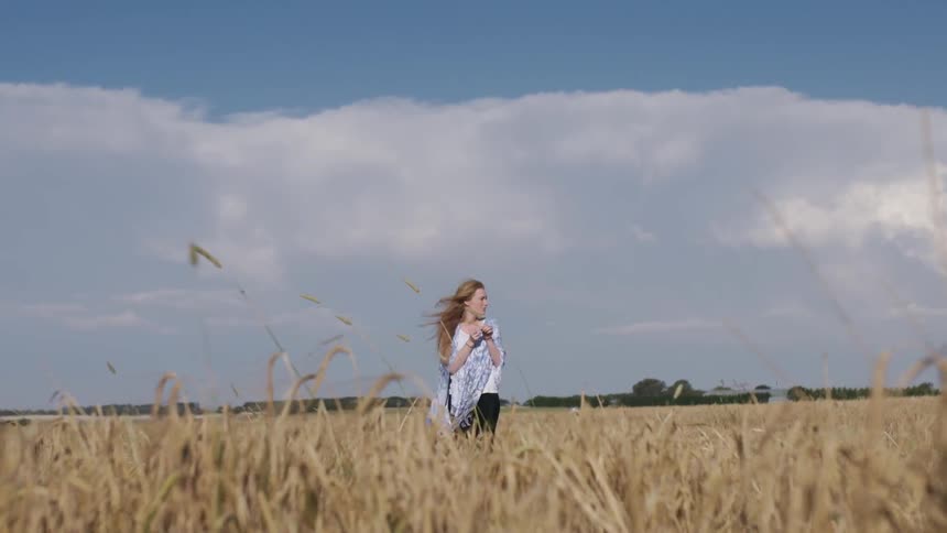 A woman is bending over to pick a stalk of grass in a field on a windy day